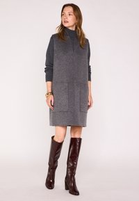 Abito grigio in misto lana con collo alto, dotato di due tasche frontali e una silhouette dritta. Abbinato a stivali in pelle bordeaux al ginocchio.