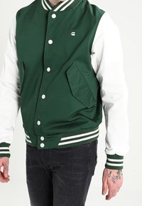 En person i en grön och vit collegejacka med framfickor och randiga manschetter, tillsammans med svarta jeans, som visar tatueringar på händerna.