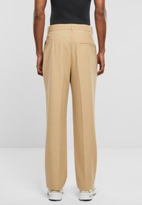 Beige Hosen mit geradem Schnitt, ausgestattet mit einer einzigen Gesäßtasche und einer glatten Textur. Kombiniert mit einem schwarzen Oberteil und weißen Sneakers.