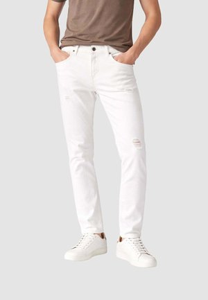 Persona che indossa jeans bianchi slim fit con dettagli usurati e sneakers bianche, mani nelle tasche, in piedi contro uno sfondo semplice.