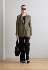 Blazer vert olive avec deux poches avant, haut noir, pantalon large noir, ballerines noires et sac tote texturé noir dans la main gauche.