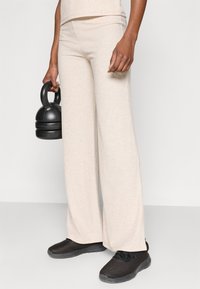 Φαρδιά μπεζ παντελόνια με ριγέ ύφανση και ψηλή μέση, συνδυασμένα με μαύρα αθλητικά παπούτσια. Το ύφασμα έχει απαλή υφή. Στο ένα χέρι κρατιέται μια βαράκλα.