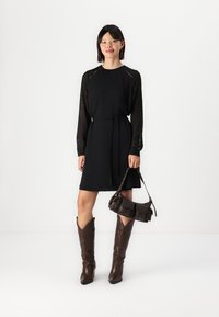 Črna obleka z dolgimi prozornimi rokavi, zategnjeno pasom, kombinirana z rjavimi kavbojskimi čevlji in majhno temno torbico. Gladek material in minimalističen dizajn.