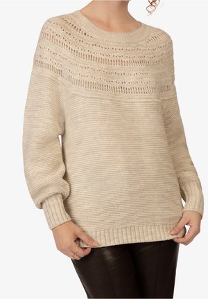 Pull beige en maille avec un motif détaillé sur le yoke, manches longues, poignets et ourlet côtelés, porté avec un pantalon noir.