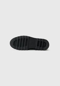 Schwarze Schuhsohle mit tiefen horizontalen Rillen und strukturierten Grip-Bereichen an der Ferse und den Zehenabschnitten vor einem schlichten Hintergrund.