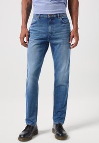 Hombre con jeans desgastados de color azul claro, zapatos de piel negros con cordones y una camiseta de color púrpura claro, de pie contra un fondo blanco liso.