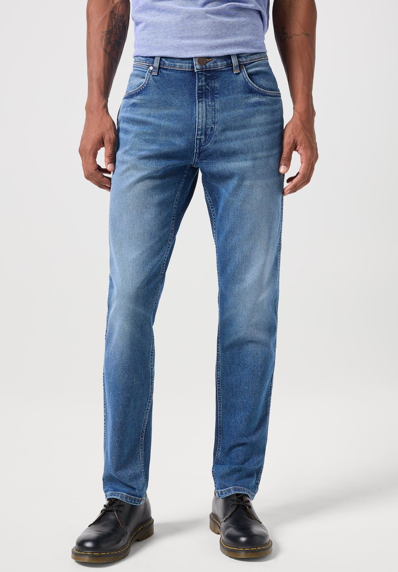 Hombre con jeans desgastados de color azul claro, zapatos de piel negros con cordones y una camiseta de color púrpura claro, de pie contra un fondo blanco liso.
