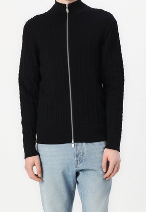 Homme portant un pull noir à torsades zippé à manches longues et un jean bleu clair, debout devant un fond blanc.