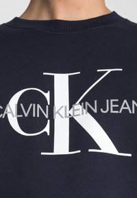 Felpa blu navy con il logo bianco "CK" in evidenza e "CALVIN KLEIN JEANS" in grigio sottostante. Ilcollo a costine aggiunge texture.