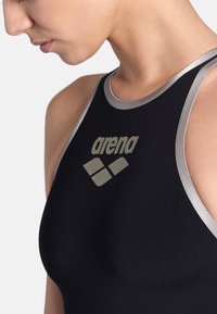 Bañador negro con acentos plateados, presentando un diseño de espalda de nadador y un destacado logo gris de "arena" en el frente. Tejido suave y elástico.