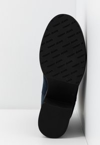 Semelle d'une botte noire avec un motif de logo "TOMMY JEANS" embossé, montrant le talon et la partie avant adhérente, sur une surface blanche d'angle.