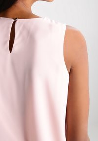Top sin mangas de color rosa, confeccionado con una tela suave, que presenta un detalle de abertura en la espalda con un pequeño broche de metal en la nuca.