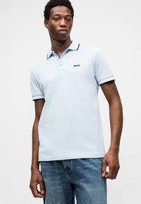 Jeune homme portant un polo bleu clair avec le logo "BOSS" et un col à bordure sombre, associé à un jean en denim bleu, debout devant un fond uni.