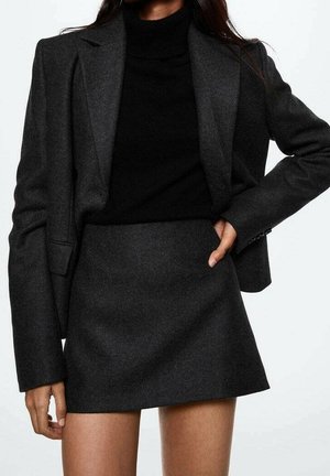 Femme portant un blazer gris foncé et une jupe assortie avec un col roulé noir, debout avec une main sur la hanche et l'autre le long du corps.