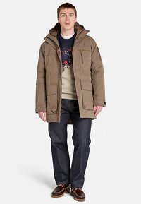 Tan puffer jacket with a hood, featuring multiple pockets and zip details, worn over a patterned sweater and dark jeans.