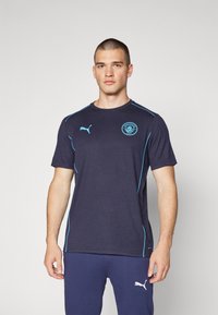 Tumši zila sporta T-kreklu ar gaiši ziliem akcentiem un Mančestras City logo uz krūtīm. Īsas piedurknes, apaļa kakla izgriezums, sportisks piegriezums.