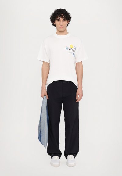 Weißes Kurzarm-T-Shirt mit mehrfarbigem floralen Stickmuster, kombiniert mit lockeren schwarzen Hosen und weißen Sneakers, während eine Jeansjacke gehalten wird.