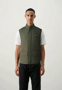 Green quilted vest with a high collar, featuring snap closure and two front pockets. Worn over a white shirt, with a logo patch on the chest.
