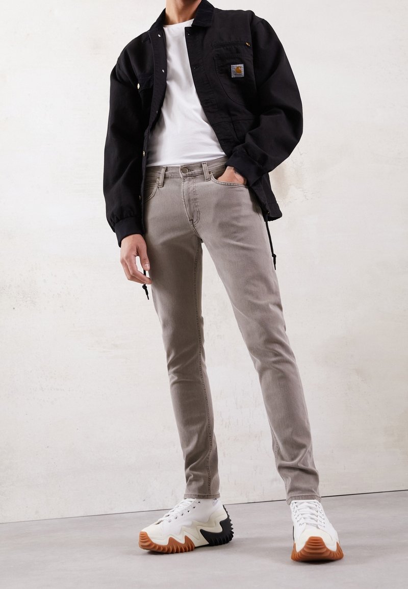 Svart jacka över en vit t-shirt, ljusgrå slim-fit jeans och vita sneakers med ett mönstrat sula. Jackan har en logotyp-patch.