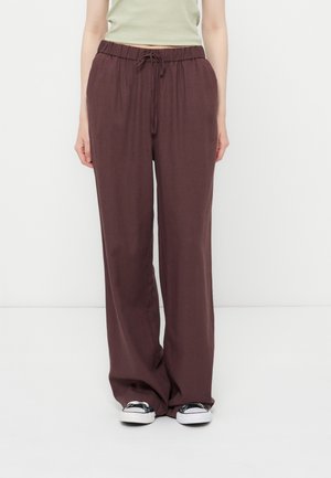 Άτομο που φοράει φαρδιά σκούρα καφέ παντελόνια με κορδόνι και ασπρόμαυρα αθλητικά παπούτσια, στέκεται μπροστά σε απλό λευκό φόντο.