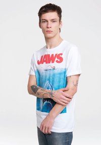Camiseta de algodón blanca con un estampado gráfico de un tiburón y un nadador con "JAWS" en letras rojas. El modelo tiene tatuajes visibles y los brazos cruzados.