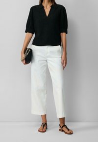 Haut noir texturé avec un col en V et des manches courtes bouffantes, associé à un pantalon blanc à jambes larges et des sandales noires à lanières ; tient une petite pochette noire.