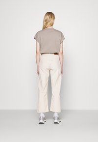 T-shirt en coton beige à manches courtes, associé à un pantalon à jambes larges de couleur claire. Des baskets grises complètent la tenue. Textures lisses visibles.