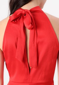 Robe sans manches rouge avec un tissu texturé, un col halter et un détail de nœud à l'arrière, créant un design de dos ouvert.