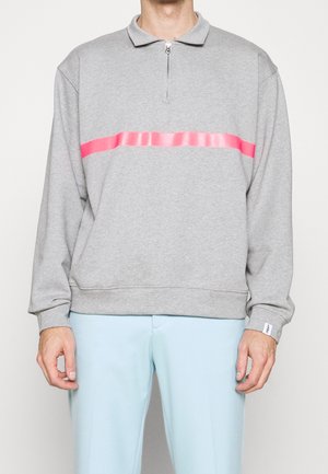 Hombre con un pulóver gris de manga larga con una franja horizontal rosa y cuello con cremallera, combinado con pantalones azul claro.