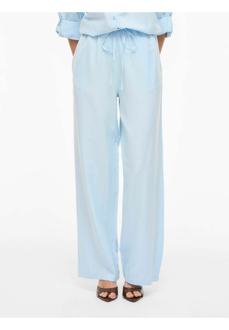 Vrouw draagt lichtblauwe broek met wijde pijpen en een trekkoord in de taille, met bruine open sandalen, staand tegen een witte achtergrond.