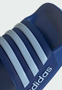 Sandal de slide azul com três riscas texturizadas, um acabamento mate suave e um logótipo elevado na alça. Textura de alta aderência na palmilha.