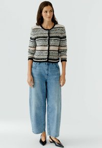 Gebreide cardigan in zwart, roze en grijs met geometrische patronen; gecombineerd met lichtblauwe, wijdvallende jeans en zwarte slingback schoenen.