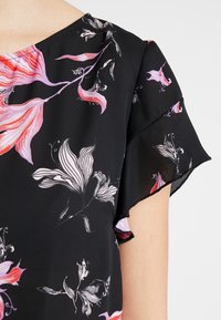 Černá blůza s květinovým potiskem v růžové a bílé barvě. Měkká, lehká látka s krátkými volánovými rukávy a kulatým výstřihem.