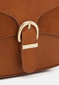 Zbliżenie na klamrę brązowej skórzanej torebki z metalowym zapięciem w kolorze złotym oraz szczegółowymi przeszyciami na skórzanym pasku i klapie.