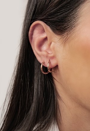 Primer plano de la oreja de una mujer con cabello largo y oscuro, que lleva dos pequeños pendientes de aro en tono oro rosa en el lóbulo de la oreja.