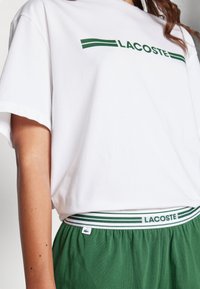 T-shirt en coton blanc avec le logo "LACOSTE" vert et des rayures, associé à un short vert avec une ceinture rayée portant le logo.