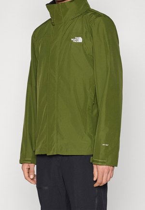 Homme portant une veste imperméable verte North Face avec un col haut et un pantalon noir, debout devant un fond uni.