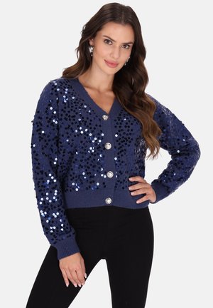 Vrouw met lang bruin haar, die een blauwe cardigan met pailletten en knopen draagt en een zwarte broek, poseert met één hand op haar heup tegen een witte achtergrond.