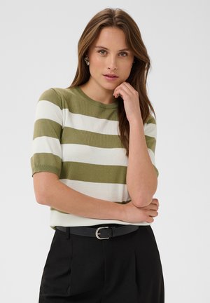 Femme aux longs cheveux bruns portant un haut à manches courtes rayé vert et blanc et un pantalon noir, posant avec une main près du menton.
