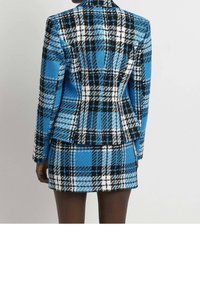 Žena oblečená v modrém, černém a bílém kostkovaném saku a odpovídající sukni, zobrazená zezadu na jednobarevném pozadí.