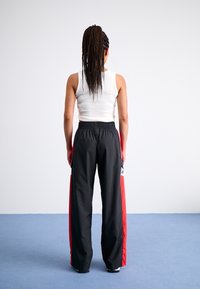 Black athletic trousers with red side accents, elastic waistband, and wide legs. Worn with a fitted white tank top. Back view.