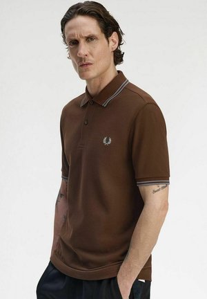 Man with short wavy hair wears a brown polo shirt with gray stripes on collar and sleeves, standing with one hand in pocket against plain background.