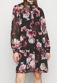 Robe noire avec un motif floral en rose et blanc, en tissu transparent. Elle possède des manches longues, un col volanté et une fermeture à boutons.