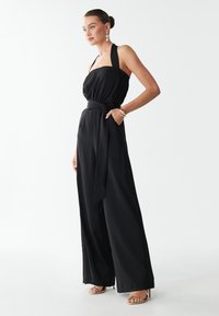 Woman wearing a black halter-neck jumpsuit with wide legs, side pockets, a waist tie, gold bracelet, earrings, and gold open-toe heels.