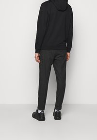 Sudadera negra con capucha y cordón, combinada con pantalones gris oscuro. Modelo con zapatillas negras con detalles blancos, visto desde atrás, mostrando el ajuste del conjunto.