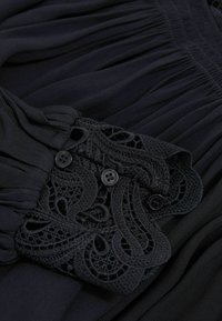 Schwarzes Chiffonkleid mit plissierter Textur und elegantem Spitzenbesatz. Verfügt über dekorative schwarze Knöpfe am Ausschnitt. Weicher, fließender Stoff.