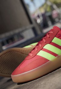 Red sneakers with neon green stripes and gum rubber soles placed on a concrete surface with an outdoor blurred background.