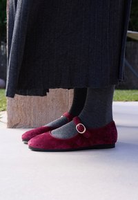 Ballerine in suede bordeaux con punta rotonda, cinturino alla caviglia con fibbia dorata, abbinate a calzini grigio scuro e gonna nera plissettata.