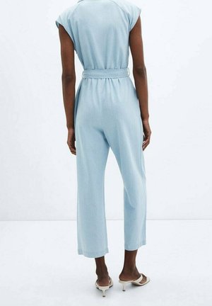 Person iført lyseblå ærmeløs jumpsuit med bælte, stående barfodet på hvid gulv mod ensfarvet hvid baggrund.