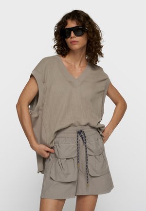 Kobieta w oversize beżowym dzianinowym topie bez rękawów, beżowych szortach z wysokim stanem i dużymi przednimi kieszeniami oraz czarnych dużych okularach przeciwsłonecznych.